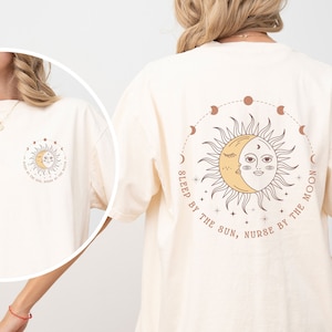 May include: Cream-colored t-shirt featuring a celestial design. The front displays a sun and moon graphic with the text "SLEEP BY THE SUN, NURSE BY THE MOON." The back of the shirt has a larger version of the design, surrounded by a circle of moon phases.