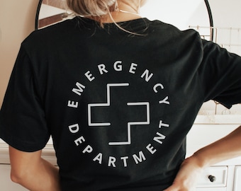 Op de afbeelding: Zwart T-shirt met een witte cirkelvormige afbeelding op de rug. De afbeelding toont een wit kruis en de tekst "Emergency Department".