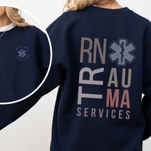 May include: A navy blue sweatshirt with the words 'RN', 'Trauma', and 'Services' printed on the back in different colors. The words are stacked vertically, with a medical symbol between 'RN' and 'Trauma'. The sweatshirt has a small, circular logo on the left chest with the words 'Trauma Services' printed inside.