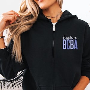 May include: Black zip-up hoodie with the name "Sophia" and the letters "BCBA" in a gradient blue color. The hoodie has a full zipper, a hood, and two front pockets. The word "BLACK" is printed at the bottom.