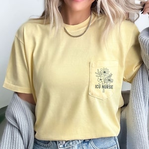 以下が含まれることがあります： 胸ポケットが付いたペールイエローの半袖Tシャツ。ポケットには、野花のブーケの黒い線画と「ICU NURSE」という文字がプリントされています。グレーのニットカーディガンとライトブルーのデニムジーンズを合わせています。