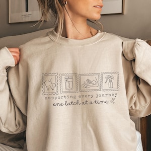 May include: Beige crewneck sweatshirt with a graphic design featuring four postage stamp-style illustrations related to breastfeeding. The text reads "supporting every journey one latch at a time" in a simple, sans-serif font. A small heart is at the end of the text.
