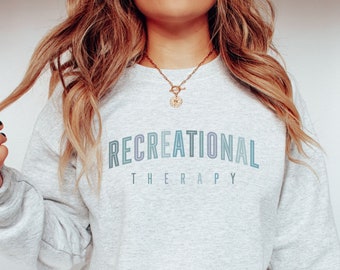 Op de afbeelding: Een lichtgrijze sweatshirt met de woorden "RECREATIONAL THERAPY" in een blauw-paarse gradiëntlettertype op de voorkant. De sweatshirt heeft een ronde hals.