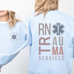 Puede incluir: Sudadera azul claro con el texto "RN" y "TRAUMA SERVICES" en rosa y gris. Una estrella m&eacute;dica gris con una serpiente est&aacute; centrada sobre el texto.