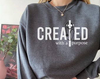 May include: Dark heather gray sweatshirt with the text "CREATED with a purpose" in white. A cross and stethoscope graphic are incorporated into the design. The sweatshirt is a crew neck style.