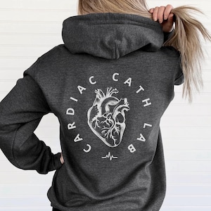 May include: Dark heather grey hoodie with a white graphic of a heart and the text "Cardiac Cath Lab" in a circle around the heart. There is a white EKG line below the heart.