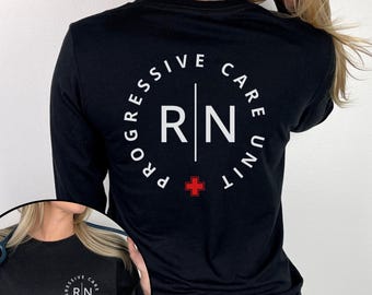 Op de afbeelding: Zwart shirt met lange mouwen en een wit cirkelvormig ontwerp op de rug. Het ontwerp bevat de woorden "PROGRESSIVE CARE UNIT" langs de rand, met "RN" en een rood kruis in het midden. Het shirt is effen zwart.