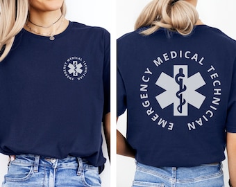 May include: A navy blue t-shirt with a white graphic of a medical symbol and the text "EMERGENCY MEDICAL TECHNICIAN". The shirt is shown on a model wearing blue jeans.