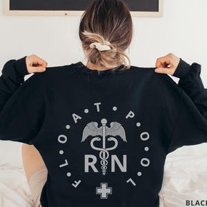 May include: Black sweatshirt with a circular design featuring the text "FLO A T POOL" and the letters "R N" with a medical symbol. A cross is below the letters. The sweatshirt is a long-sleeved crew neck.