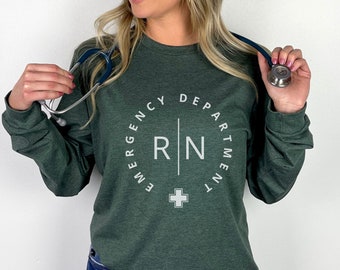 May include: A dark green long-sleeve shirt with the text "EMERGENCY DEPARTMENT RN" in a white circular design. A white cross is below the text. The shirt is made of a heather forest material.