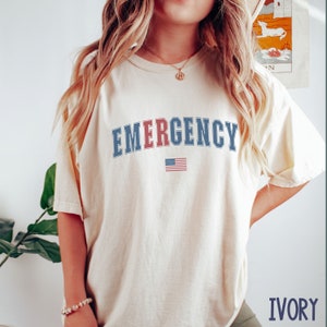 Peut inclure: Un t-shirt à manches courtes de couleur ivoire avec le mot EMERGENCY imprimé dans une police universitaire sur la poitrine. Les lettres sont principalement bleues, avec la lettre R en rouge. Sous le texte se trouve un petit drapeau américain.