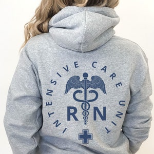 Peut inclure: Sweat-shirt &agrave; capuche gris avec un motif bleu repr&eacute;sentant un symbole m&eacute;dical, un serpent enroul&eacute; autour d'un b&acirc;ton avec des ailes, et les lettres "RN" dans un cercle. Le texte "INTENSIVE CARE UNIT" entoure le symbole.