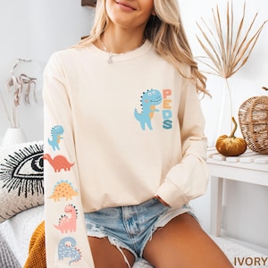May include: Ivory long-sleeve shirt with a blue dinosaur graphic and the word "PEDS". The left sleeve features five colorful dinosaur illustrations. The shirt is made of a soft material.