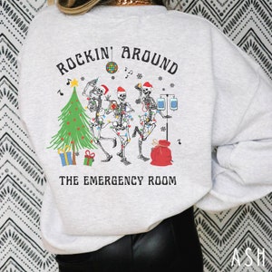 Op de afbeelding: Een lichtgrijs sweatshirt met ronde hals en een afbeelding van drie skeletten met kerstmutsen die dansen rond een kerstboom met medische apparatuur, een discobal en de tekst "ROCKIN' AROUND THE EMERGENCY ROOM" in zwarte letters.
