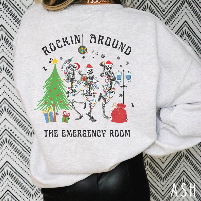 Op de afbeelding: Een lichtgrijs sweatshirt met ronde hals en een afbeelding van drie skeletten met kerstmutsen die dansen rond een kerstboom met medische apparatuur, een discobal en de tekst "ROCKIN' AROUND THE EMERGENCY ROOM" in zwarte letters.
