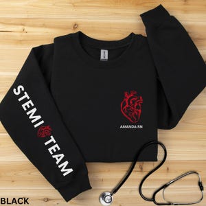 May include: A black long-sleeve sweatshirt featuring a red anatomical heart graphic on the left chest with the text AMANDA RN below it. The left sleeve displays the white text STEMI TEAM with a small red anatomical heart icon between the words.