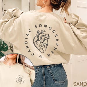 May include: Sand-colored sweatshirt with the text "CARDIAC SONOGRAPHER" encircling a detailed illustration of a heart. Below the heart is a graphic of a heartbeat. The sweatshirt is a neutral color and has a relaxed fit.