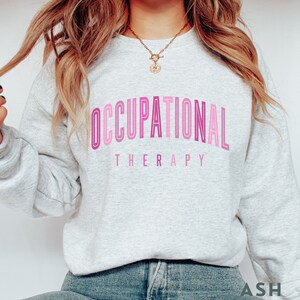 Op de afbeelding: Een lichtgrijs sweatshirt met ronde hals en de tekst OCCUPATIONAL THERAPY in een verloop van roze en magentatinten. Het woord OCCUPATIONAL is gebogen in een vet, gelaagd lettertype, terwijl THERAPY in een eenvoudig schreefloos lettertype is geschreven.