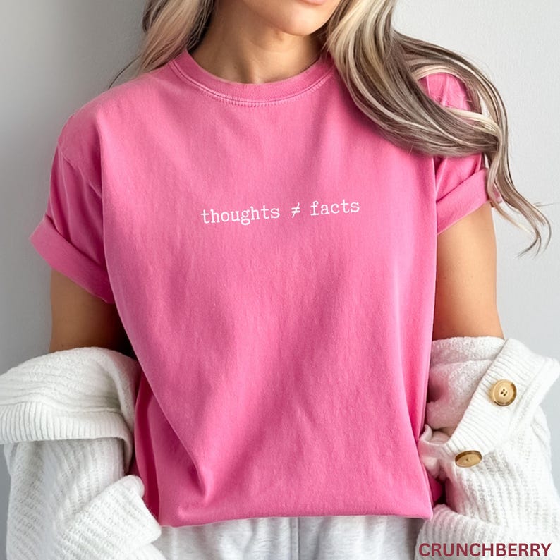Puede incluir: Camiseta rosa brillante con la frase "thoughts &ne; facts" en blanco. La camiseta de manga corta, de tejido suave, tiene las mangas dobladas. Forma parte de una colecci&oacute;n de ropa.