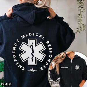 May include: Black zip-up hoodie featuring a white graphic on the back with the words "Emergency Medical Responder" encircling a white Star of Life symbol. The lower left corner of the hoodie says "BLACK". A smaller image shows the front of the hoodie with the text "Elizabeth RN, BSN".