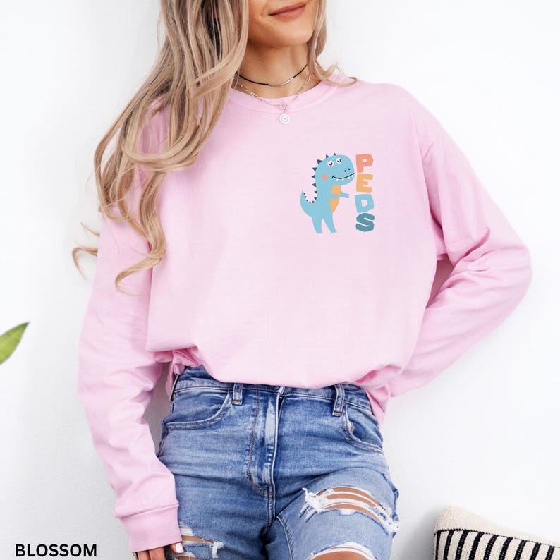 May include: A light pink long-sleeve sweatshirt with a blue dinosaur graphic and the word "PEDS" in orange, green, and blue. The sweatshirt is made of a soft material and is perfect for everyday wear.