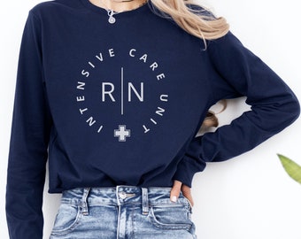 Puede incluir: Una camiseta azul marino de manga larga con un gráfico circular blanco en el pecho. El texto dice INTENSIVE CARE UNIT con las letras RN separadas por una línea vertical y un pequeño símbolo de cruz situado en la parte inferior del círculo.