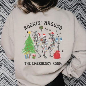 Op de afbeelding: Een beige sweater met ronde hals en een grafische print van drie skeletten met kerstmutsen, dansend rond een kerstboom met cadeautjes, een discobal en medische apparatuur, met de tekst ROCKIN' AROUND THE EMERGENCY ROOM.