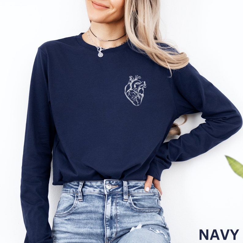 Puede incluir: Una camiseta de manga larga azul marino con un gr&aacute;fico de coraz&oacute;n anat&oacute;mico blanco impreso en la parte superior izquierda del pecho. La prenda tiene cuello redondo y mangas largas con pu&ntilde;os acanalados, combinada con vaqueros azules desgastados.