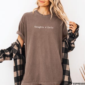 Puede incluir: Camiseta color espresso con el texto blanco "thoughts &ne; facts". La camiseta tiene las mangas enrolladas y se combina con una camisa de franela a cuadros en negro y beige. El estilo general es informal y c&oacute;modo.