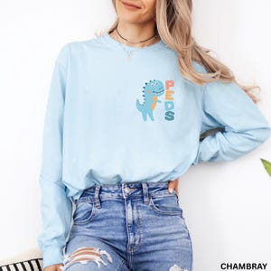 May include: A light blue long-sleeve shirt with a dinosaur graphic and the word "PEDS". The dinosaur is blue with orange accents. The shirt is made of a soft, comfortable material.