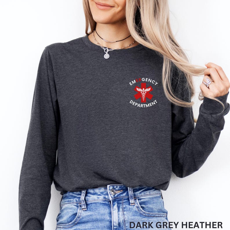 Puede incluir: Una camiseta de manga larga en color gris jaspeado oscuro con un emblema rojo de la Estrella de la Vida en el pecho izquierdo. El emblema incluye un caduceo blanco y el texto EMERGENCY DEPARTMENT impreso en blanco alrededor de la estrella.