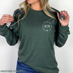 May include: A dark heather forest green long-sleeve shirt featuring a white circular graphic on the left chest. The text inside the circle reads RN and Neonatal Intensive Care Unit. A silver and blue stethoscope is draped around the neck area.