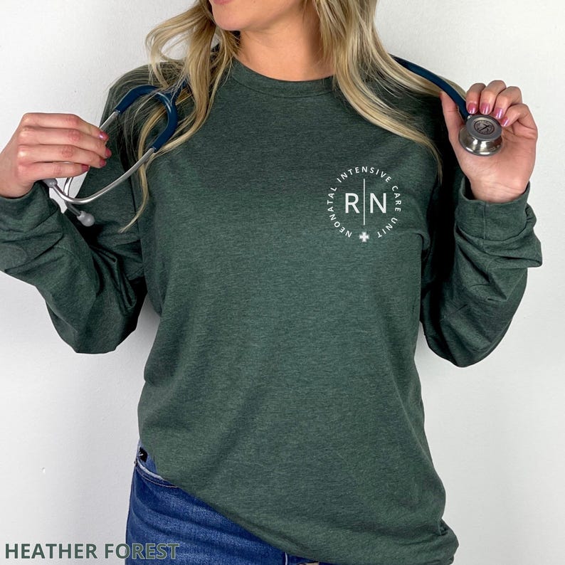 May include: A dark heather forest green long-sleeve shirt featuring a white circular graphic on the left chest. The text inside the circle reads RN and Neonatal Intensive Care Unit. A silver and blue stethoscope is draped around the neck area.