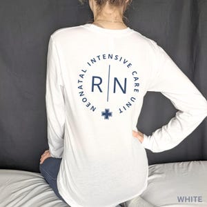 Puede incluir: Camiseta blanca de manga larga con un gr&aacute;fico circular azul que dice "Neonatal Intensive Care Unit RN" con una cruz en el centro.