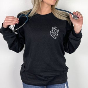 Puede incluir: Una camiseta negra de manga larga con una ilustraci&oacute;n blanca de un coraz&oacute;n anat&oacute;mico en la parte superior izquierda del pecho. La prenda se combina con un estetoscopio azul oscuro alrededor del cuello, mostrando un dise&ntilde;o m&eacute;dico.