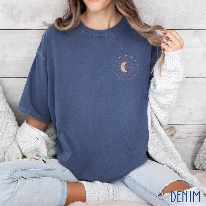 May include: A denim blue t-shirt with a crew neck and short sleeves. The shirt features a celestial design on the upper left chest, including a sun, moon, and stars. The word "DENIM" is printed at the bottom right.