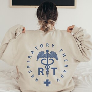 May include: A beige sweatshirt with a blue graphic on the back. The graphic is a circle with the words "Respiratory Therapy" around the outside. Inside the circle is a medical symbol with a snake wrapped around a staff, a cross at the bottom, and the letters "RT" in the center.