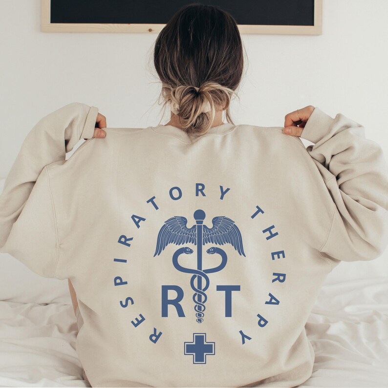 May include: A beige sweatshirt with a blue graphic on the back. The graphic is a circle with the words "Respiratory Therapy" around the outside. Inside the circle is a medical symbol with a snake wrapped around a staff, a cross at the bottom, and the letters "RT" in the center.