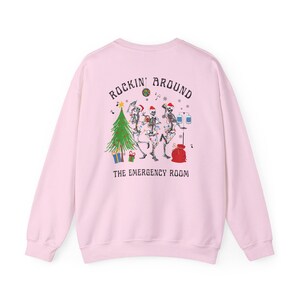 Op de afbeelding: Een lichtroze sweater met ronde hals en een afbeelding op de achterkant van drie skeletten met kerstmutsen die dansen rond een kerstboom met een discobal, cadeautjes en een infuuspaal. De tekst luidt Rockin' Around The Emergency Room.