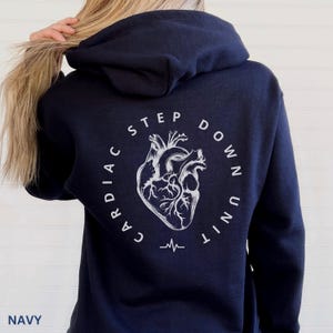 Op de afbeelding: Marineblauwe hoodie met een witte afbeelding van een hart en de woorden "CARDIAC STEP DOWN UNIT" in een cirkelvormig ontwerp. Het woord "NAVY" staat onderaan gedrukt. Onder de hartafbeelding staat een hartslaglijn.