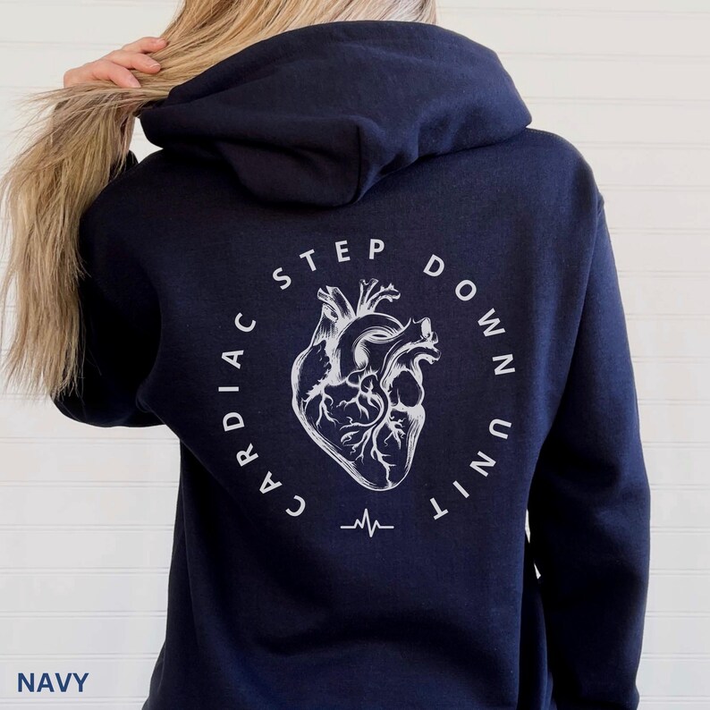 Op de afbeelding: Marineblauwe hoodie met een witte afbeelding van een hart en de woorden "CARDIAC STEP DOWN UNIT" in een cirkelvormig ontwerp. Het woord "NAVY" staat onderaan gedrukt. Onder de hartafbeelding staat een hartslaglijn.