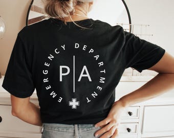 May include: Black t-shirt with a white circular design on the back that reads "EMERGENCY DEPARTMENT" around the top and sides. Inside the circle are the letters "P A" separated by a vertical line, with a white cross below. The word "BLACK" is printed in white at the bottom right.