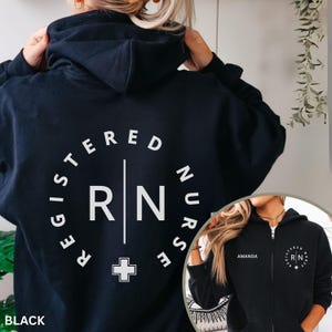May include: A black zip-up hoodie featuring a white circular graphic on the back with the text REGISTERED NURSE and RN separated by a vertical line and a cross symbol. The front displays the name AMANDA and a smaller version of the circular logo.