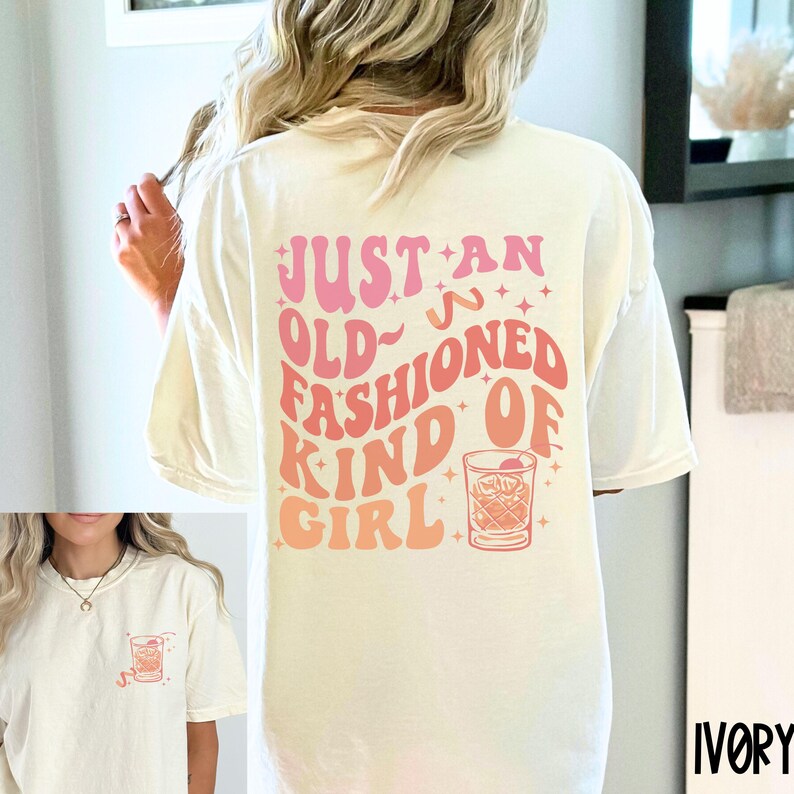 Peut inclure: Un t-shirt blanc avec le texte "Just an old-fashioned kind of girl" en lettres roses et orange. Une illustration vintage d'un verre &agrave; cocktail avec une cerise se trouve &agrave; droite du texte.