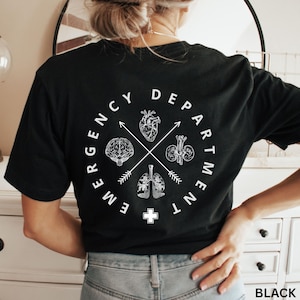 Op de afbeelding: Zwart T-shirt met een wit grafisch ontwerp. Het ontwerp toont een cirkelvormig embleem met de woorden "EMERGENCY DEPARTMENT" en illustraties van de hersenen, het hart, de nieren en de longen. Onderaan staat een wit kruis.