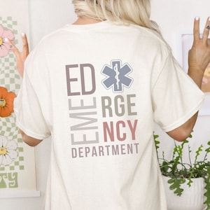 May include: A cream-colored t-shirt featuring a graphic on the back with the text ED, RGE, and NCY arranged vertically alongside the word DEPARTMENT. A blue Star of Life symbol with a serpent and staff is positioned next to the letters ED.
