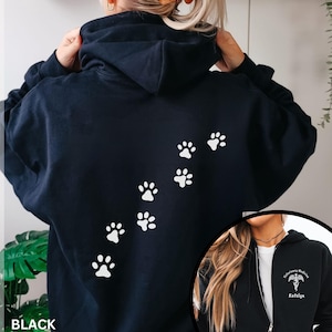 Könnte beinhalten: Ein schwarzer Kapuzenpullover mit Reißverschluss und einer weißen Pfotenabdruck-Spur auf der Rückseite. Auf der linken Brust befindet sich ein weißes Caduceus-Symbol mit dem Text Veterinary Medicine und dem Namen Katelyn in weißer Schrift.