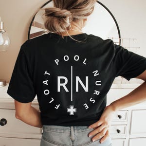 May include: Black t-shirt featuring a white circular design on the back. The design includes the words "POOL FLOAT NURSE" and "RN" with a cross symbol. The shirt is a crew neck and has short sleeves.
