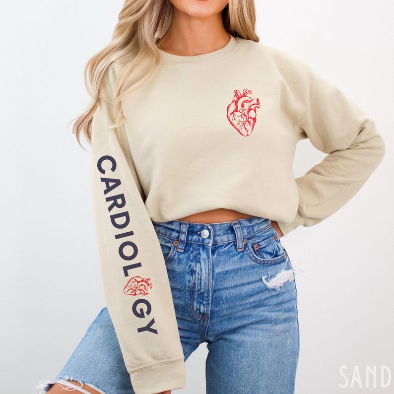 Peut inclure: Un sweat-shirt beige court avec le mot "CARDIOLOGY" imprim&eacute; verticalement sur la manche gauche. Un c&oelig;ur anatomique rouge est imprim&eacute; sur le devant du sweat-shirt.