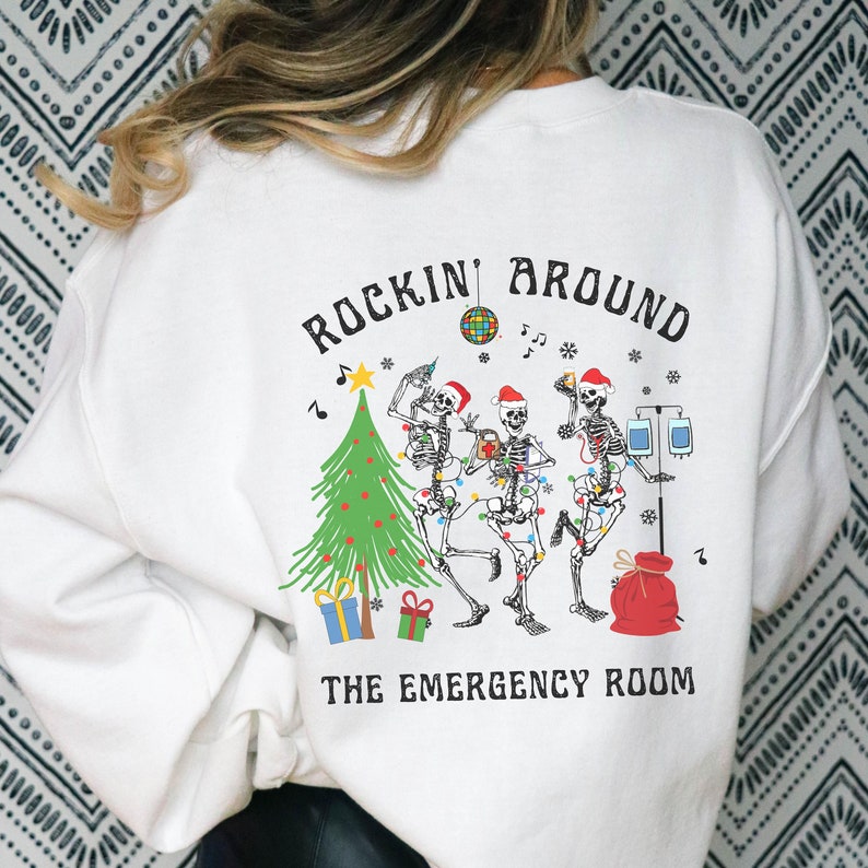 Op de afbeelding: Een wit sweatshirt met een afbeelding op de achterkant van drie skeletten met kerstmutsen, een kerstboom, cadeaus, een discobal en medische apparatuur. De tekst "ROCKIN' AROUND THE EMERGENCY ROOM" staat in zwarte letters op het kledingstuk.